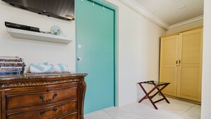 Una habitación con una puerta verde azulada, un aparador de madera con cajones, una mesita con un ventilador y un armario amarillo.