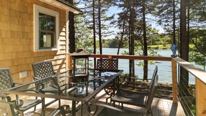 A deck with glass-top tables and chairs, overlooking a lake with tall trees. - Waterfront Cottage in Maine | Seegar Camp, Sunny 2 (Tenants Harbor)