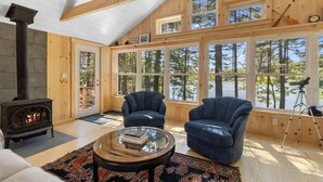 A cozy cabin interior with a wood-burning stove, two blue armchairs, a glass coffee table, and large windows offering a view of the forest. - Waterfront Cottage in Maine | Seegar Camp, Sunny 2 (Tenants Harbor)