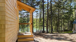 A wooden cabin with a porch, a gravel path, and a forested area with a lake in the background. - Lindquist Cottage | Waterfront Studio with King Be (Tenants Harbor)