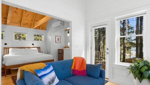 A room with a bed, a blue sofa, a large window, and a view of trees. - Lindquist Cottage | Waterfront Studio with King Be (Tenants Harbor)