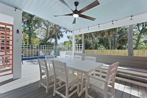 Alpendre coberto com mesa e cadeiras, ventoinha no teto e vista para uma piscina e árvores.