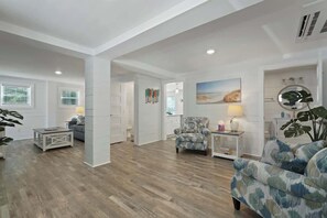 Uma sala de estar ampla com piso de madeira, uma poltrona estampada, uma mesa pequena e um quadro na parede.