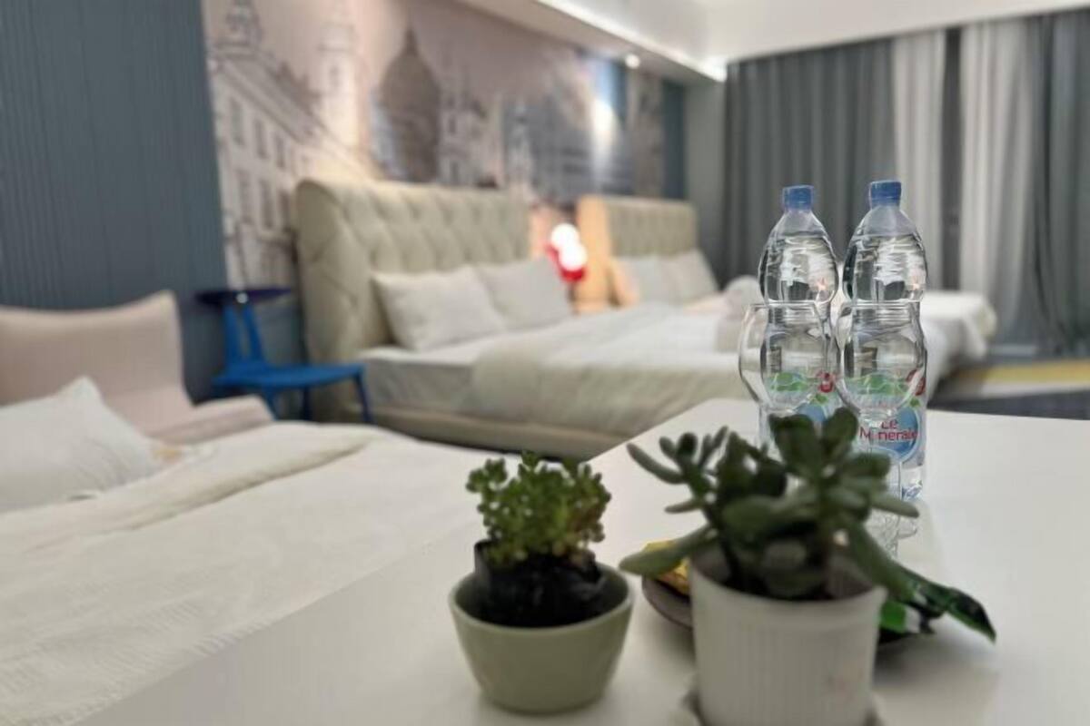 Quarto de hotel com uma cama, duas plantas em vasos e duas garrafas de água no criado-mudo.