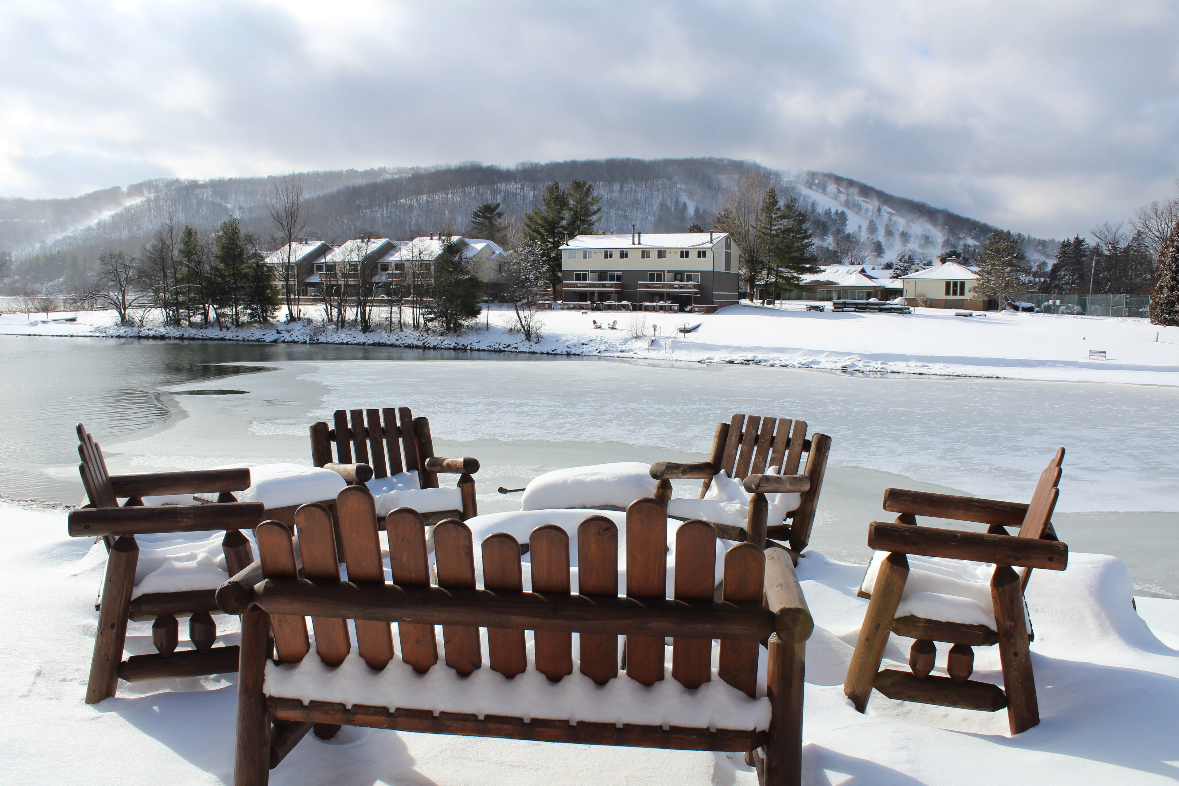 A wooden bench and chairs covered in snow, a snow-covered landscape, and buildings in the background.