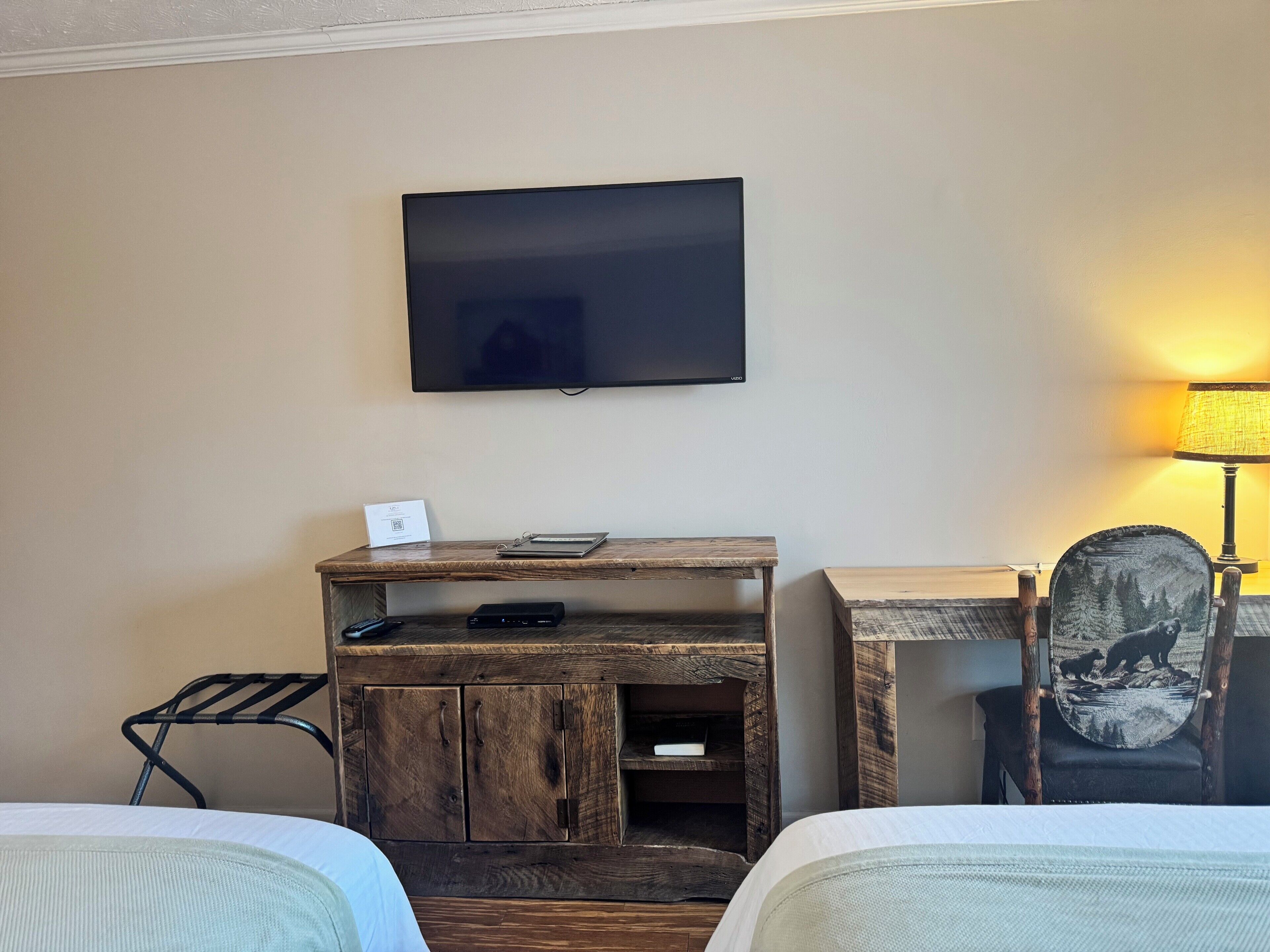 A hotel room with a bed, a desk with a television, a lamp, and a chair with a forest design.