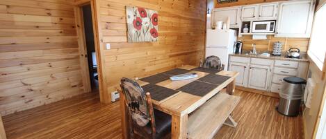 A wooden interior with a dining table, chairs, a bench, a trash can, and a kitchen area with cabinets and appliances.