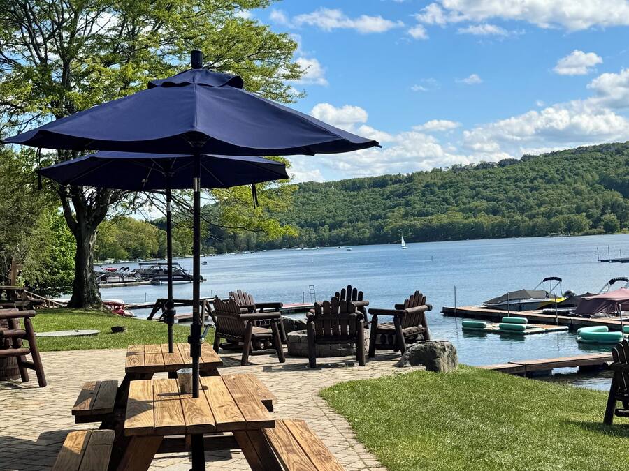 A lakeside picnic area with wooden benches and a table under a blue umbrella, overlooking a lake with boats and a forested hill in the background.