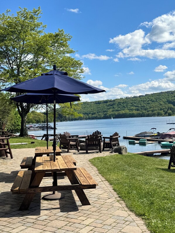 Un espace de pique-nique au bord du lac, avec des bancs en bois et une table sous un parasol bleu, d’où l’on peut admirer le lac, les bateaux qui y naviguent et une colline boisée en arrière-plan.