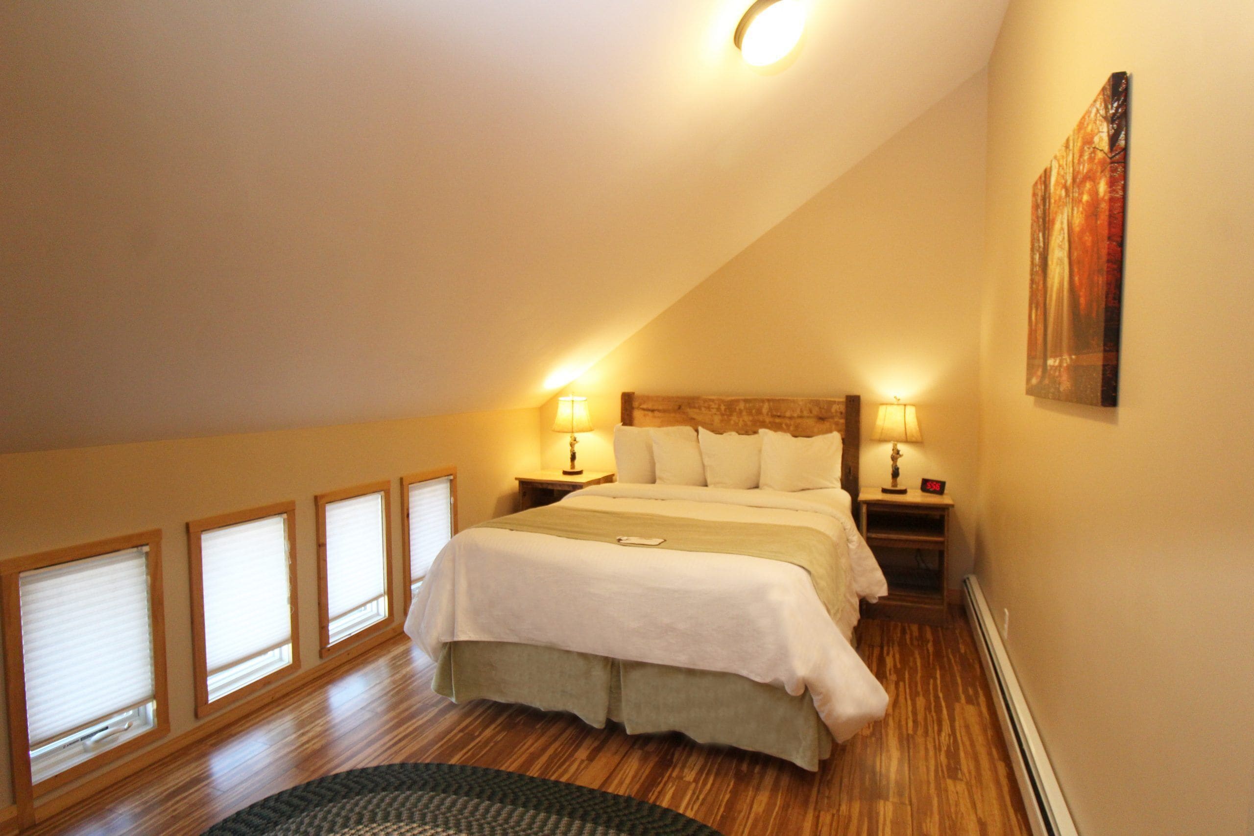 A bedroom with a large bed, two bedside tables with lamps, a window with blinds, and a painting on the wall.
