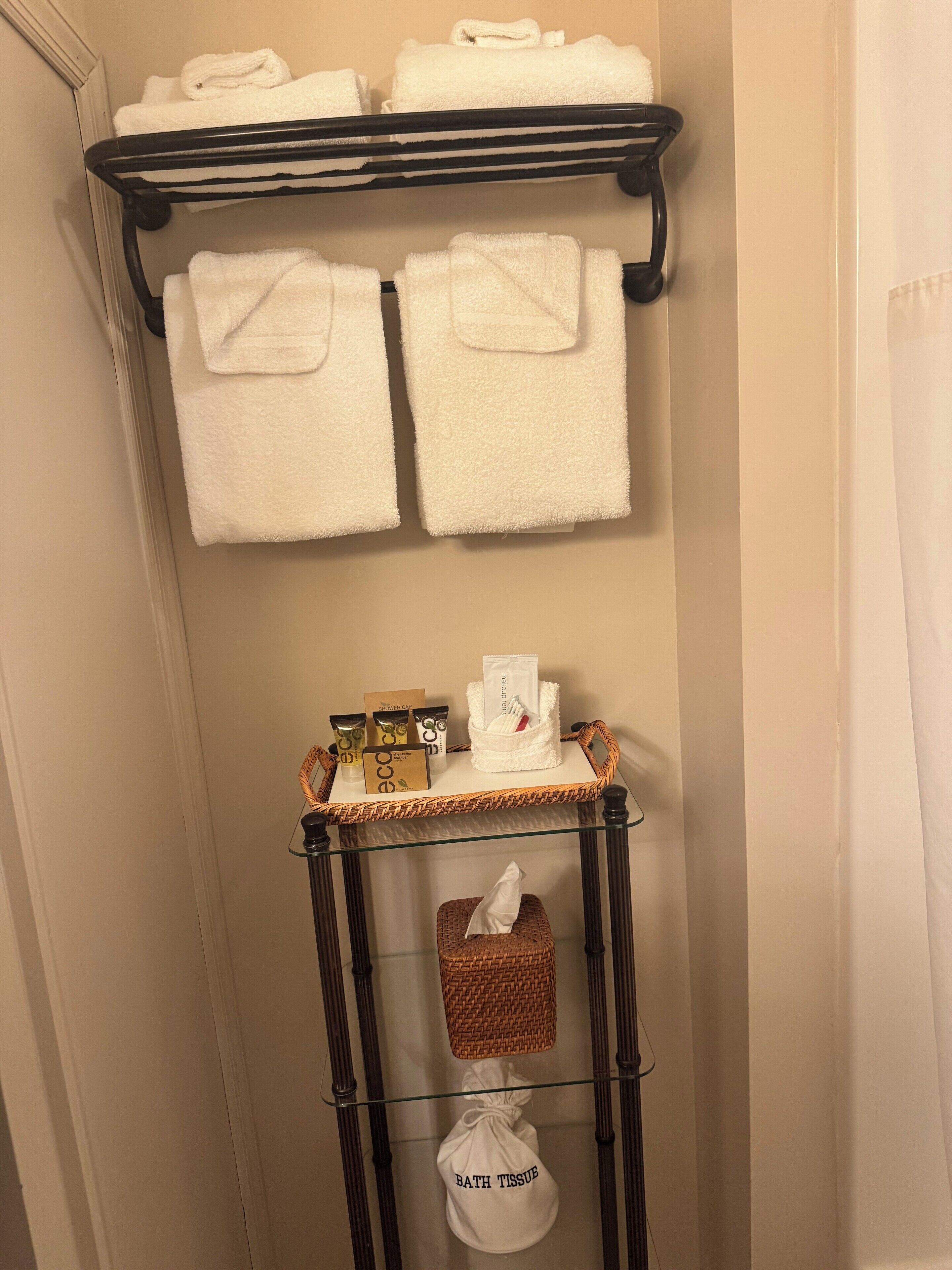 A bathroom with a towel rack holding two towels, a small table with toiletries, and a basket labeled 'bath tissue'.