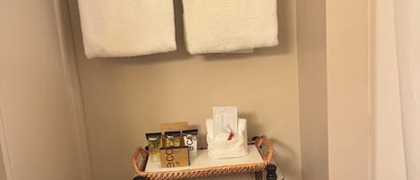 A bathroom with a towel rack holding two towels, a small table with toiletries, and a basket labeled 'bath tissue'.