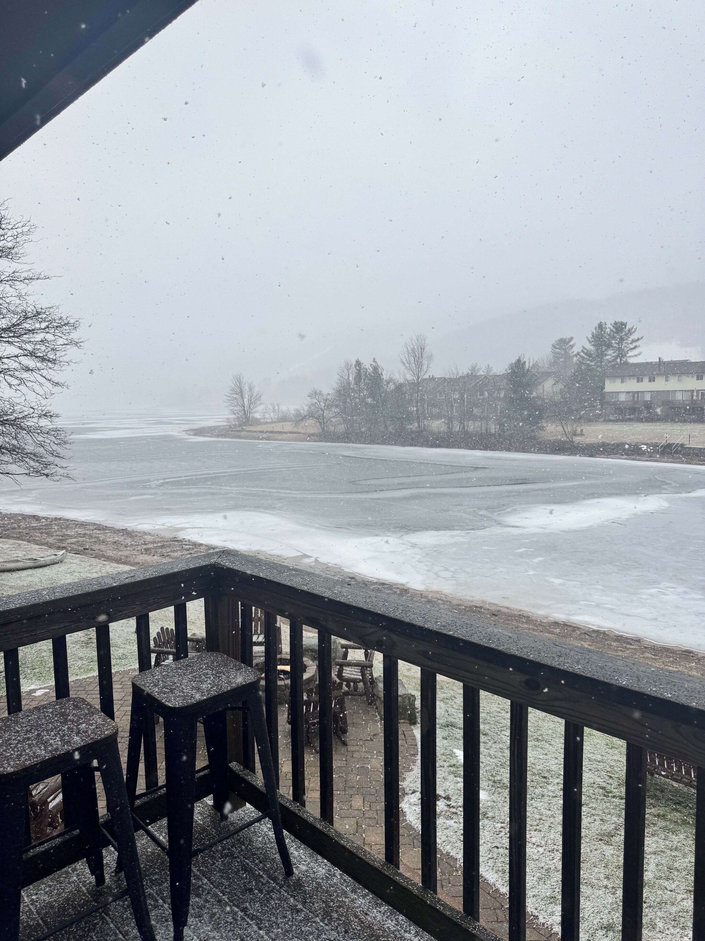 A snowy outdoor area with a wooden deck, chairs, and a view of a frozen body of water and buildings in the background.