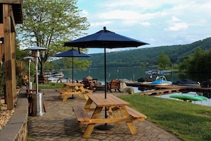 An outdoor seating area with wooden picnic tables and blue umbrellas, overlooking a lake with boats docked.