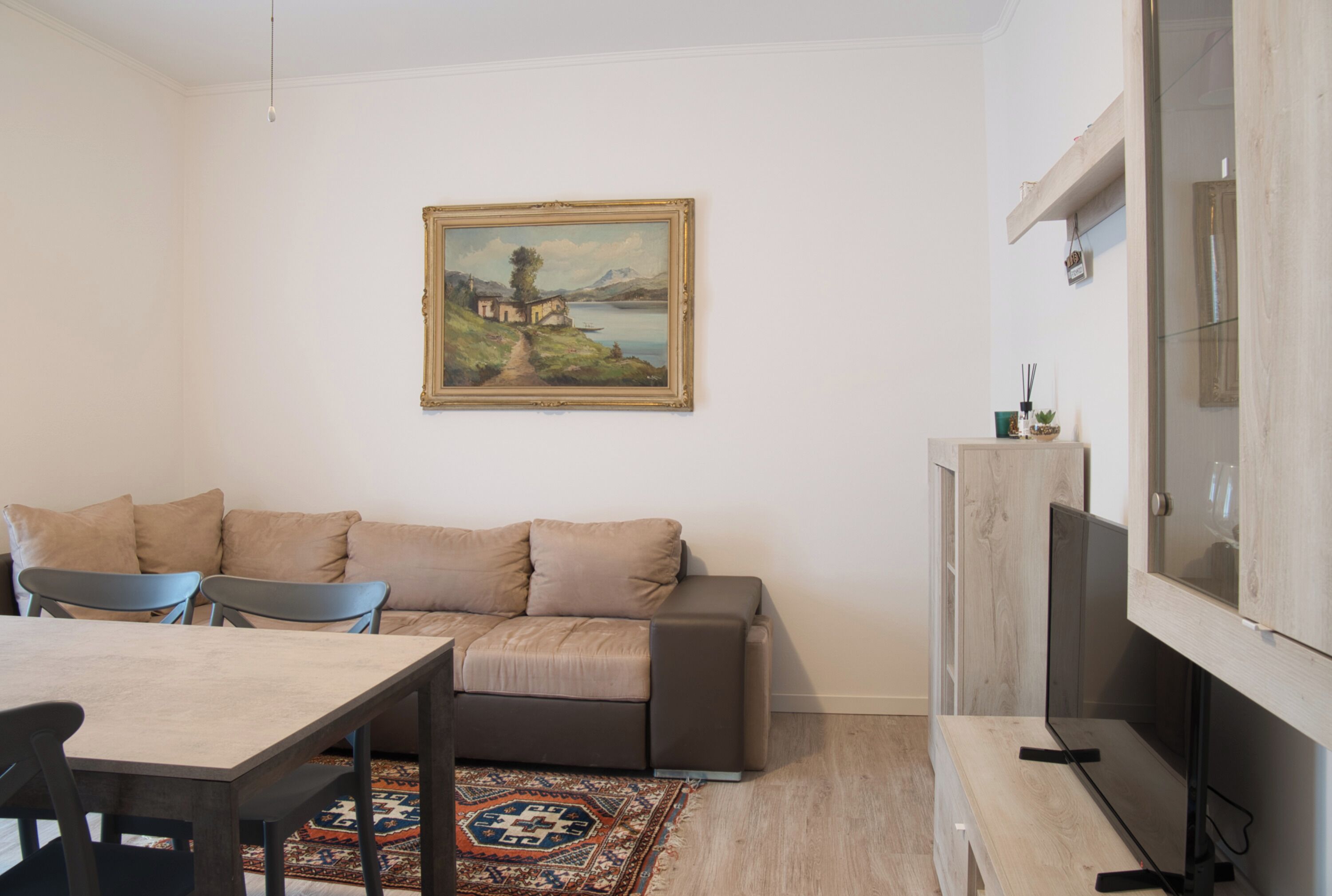 A modern living room with a sofa, a dining table, and chairs. There is a painting on the wall and a TV on a wooden stand.