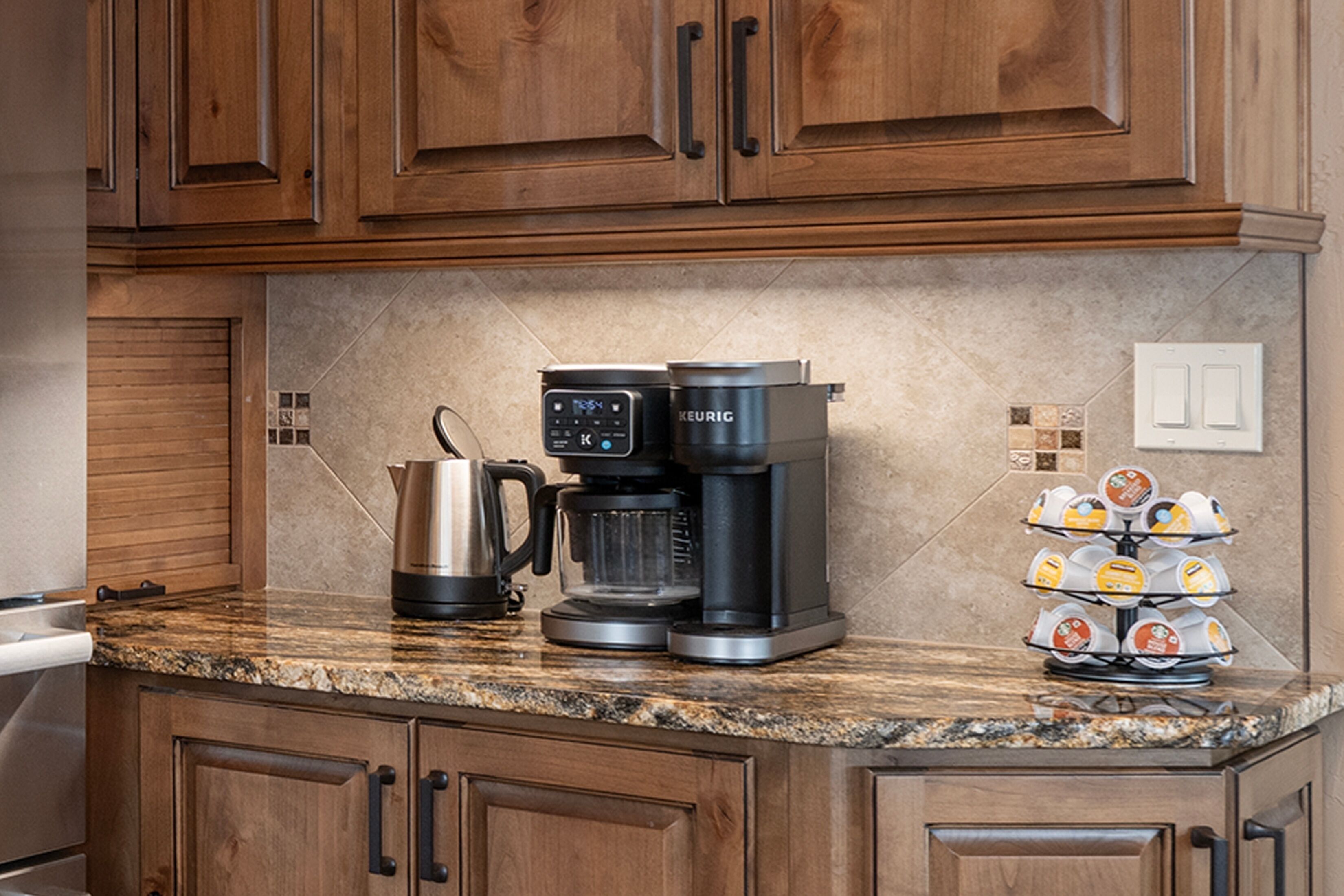 Una cocina con armarios de madera, cafetera, hervidor y soporte para cápsulas de café.