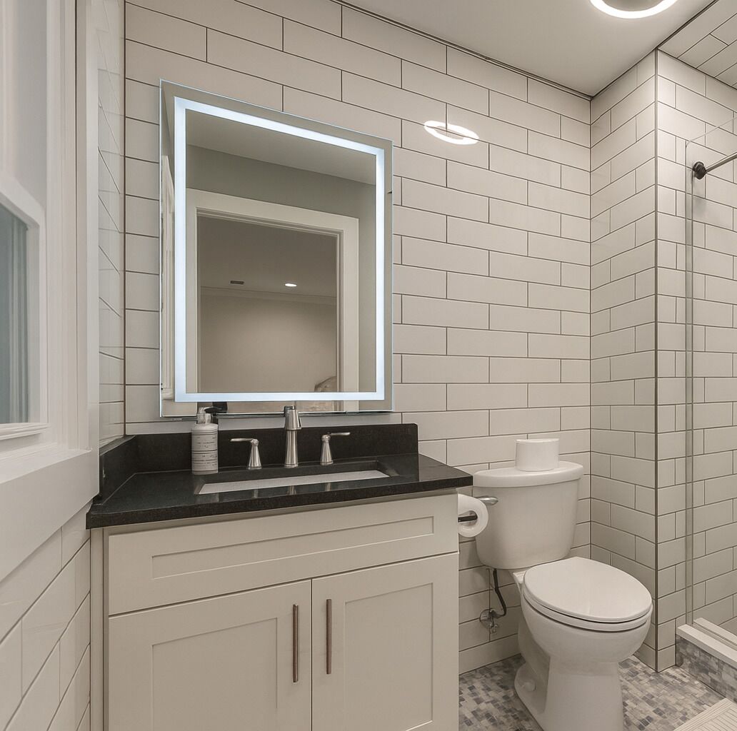 A bathroom with a white tiled wall, a black countertop, a white toilet, and a mirror with a lighted frame.