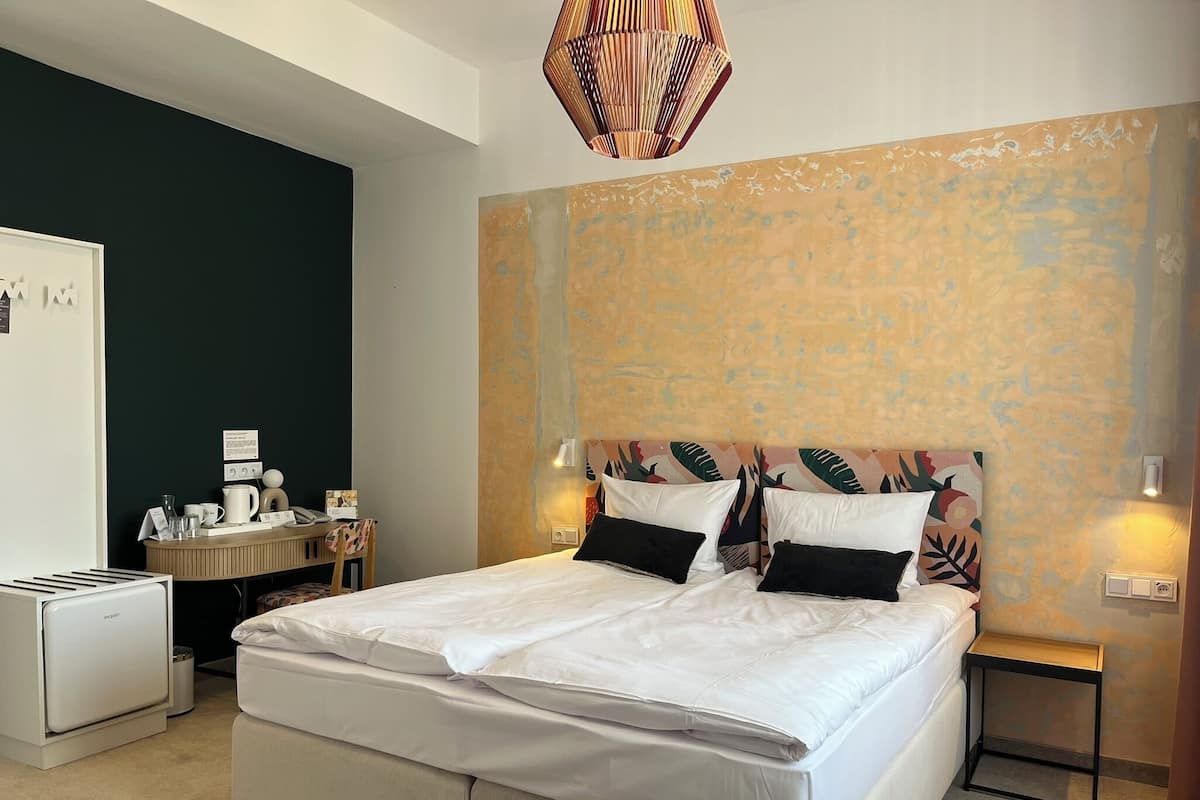 Habitación de hotel con cama, mesita de noche, lámpara, una pequeña mesa con tetera y un refrigerador.