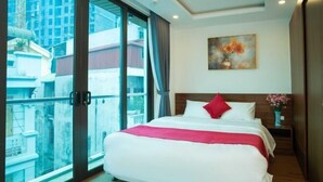 Habitación de hotel con una cama grande, vista de la ciudad a través de un ventanal y un cuadro en la pared, además de un florero con flores.