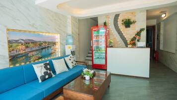 Un hall de hotel con un sofá azul, un mostrador de recepción, una máquina expendedora de bebidas roja y un cuadro grande con una vista de la ciudad.