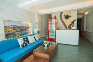 A hotel lobby with a blue sofa, a reception desk, a red beverage machine, and a large framed picture of a cityscape. - Atypik Vietnam House (Hanoi)