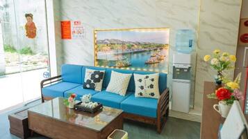 Habitación de hotel con sofá azul, mesa de centro de madera, dispensador de agua y un cuadro con una vista panorámica.