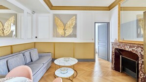 Una habitación con un sofá gris, una mesita redonda pequeña y una chimenea con un espejo de marco dorado.