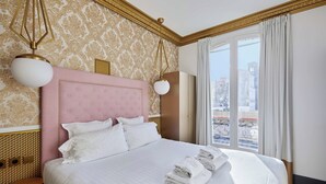 Una habitación de hotel con una cama grande, un cabecero rosa y vistas a la ciudad a través de un ventanal amplio.