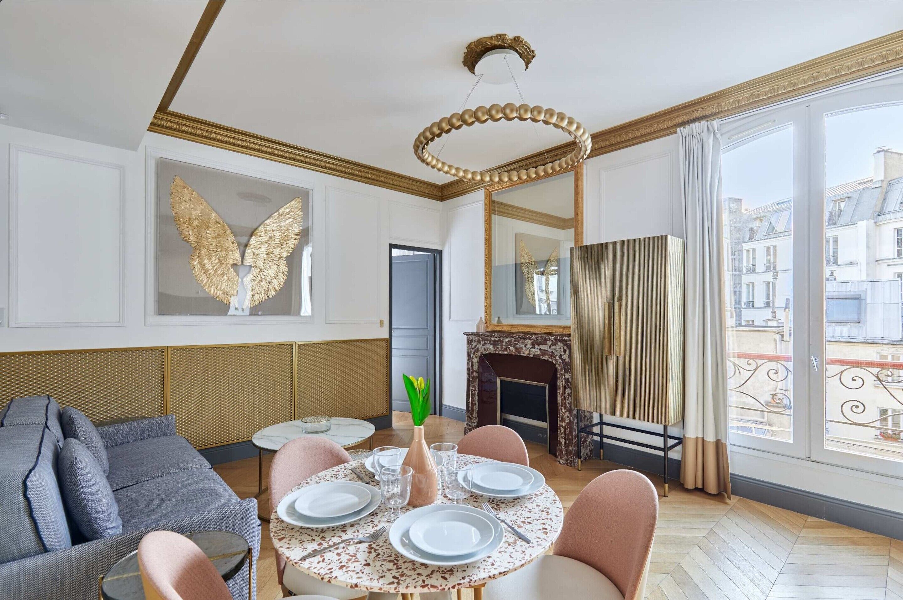Un salon comprenant une table de salle à manger ronde, préparée pour quatre personnes, un canapé, une cheminée et une grande fenêtre donnant sur des immeubles.