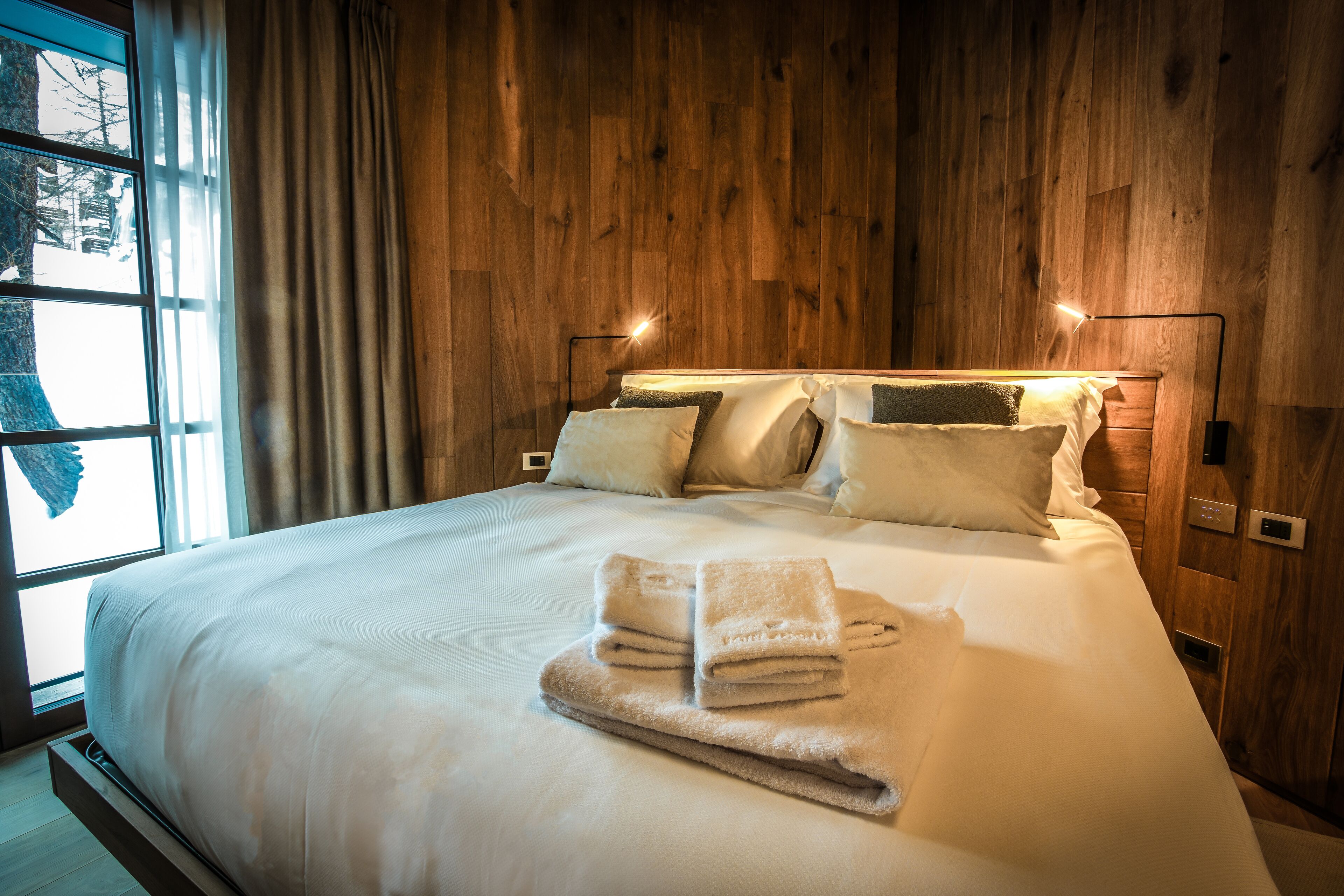 Een keurig opgemaakt bed met een houten hoofdbord, een bedlampje en een raam met gordijnen.