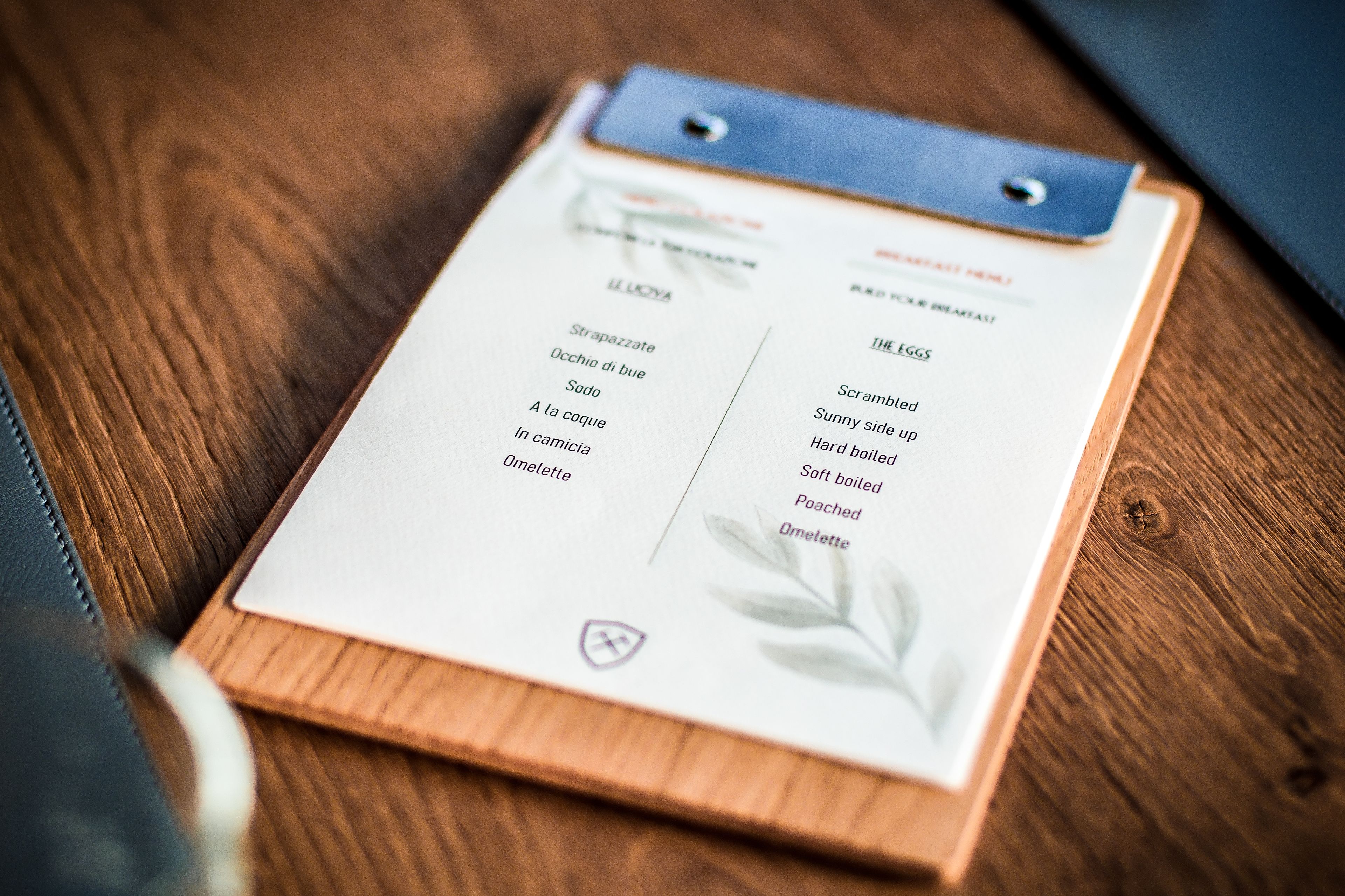 A menu on a wooden clipboard with a leather strap, listing breakfast items like scrambled eggs and omelettes.