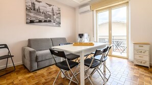 Uma sala de estar moderna com um sofá cinzento, uma mesa de jantar branca com quatro cadeiras, uma pintura em preto e branco da paisagem urbana na parede e uma varanda com vista.