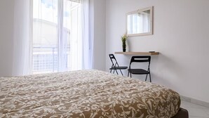 Quarto com uma cama grande, uma escrivaninha com duas cadeiras, um espelho e uma janela com cortinas leves.