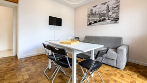 Uma sala de estar moderna com uma mesa branca, quatro cadeiras pretas, um sofá cinza, uma TV de tela plana na parede e uma pintura de paisagem urbana em preto e branco.