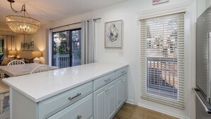 Una cocina con una isla grande, armarios blancos y una lámpara de araña. Hay un comedor con una mesa y sillas, y una ventana con persianas.