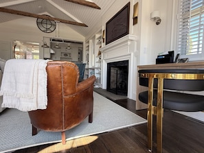 A living room with a fireplace, a brown leather chair, a wooden table, and a kitchen in the background. - Watch Hill Luxury by the Sea, Iconic 1852 School House, Beside Ocean House Hotel (Westerly)