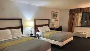 A hotel room with two beds, a nightstand with a lamp, a desk, a chair, a window with curtains, and an air conditioning unit. - Neat and Clean Rooms that are Affordable! (Hagerstown)
