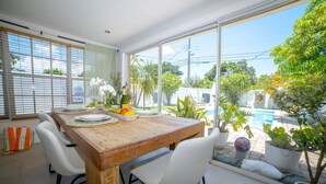 A dining area with a wooden table, white chairs, and a view of a swimming pool and greenery. - Chic MangoTree villa/Heated Pool (Fort Lauderdale)