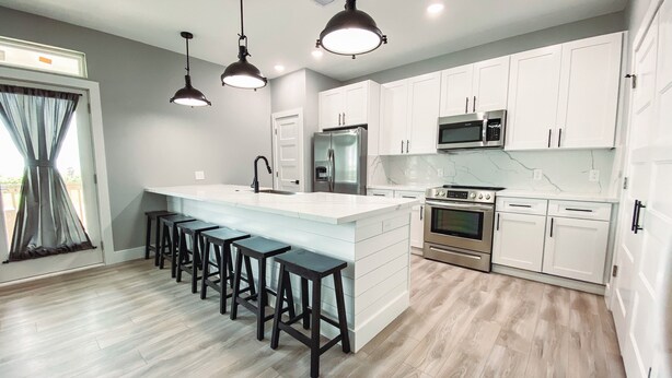 A modern kitchen with a central island, bar stools, white cabinets, stainless steel appliances, and pendant lighting. - Charming 3-bedroom, pet friendly, house in Crystal Beach. (Bolivar Peninsula)