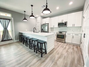 Uma cozinha moderna com uma ilha central, banquetas de bar, armários brancos, eletrodomésticos de aço inoxidável e iluminação pendente.