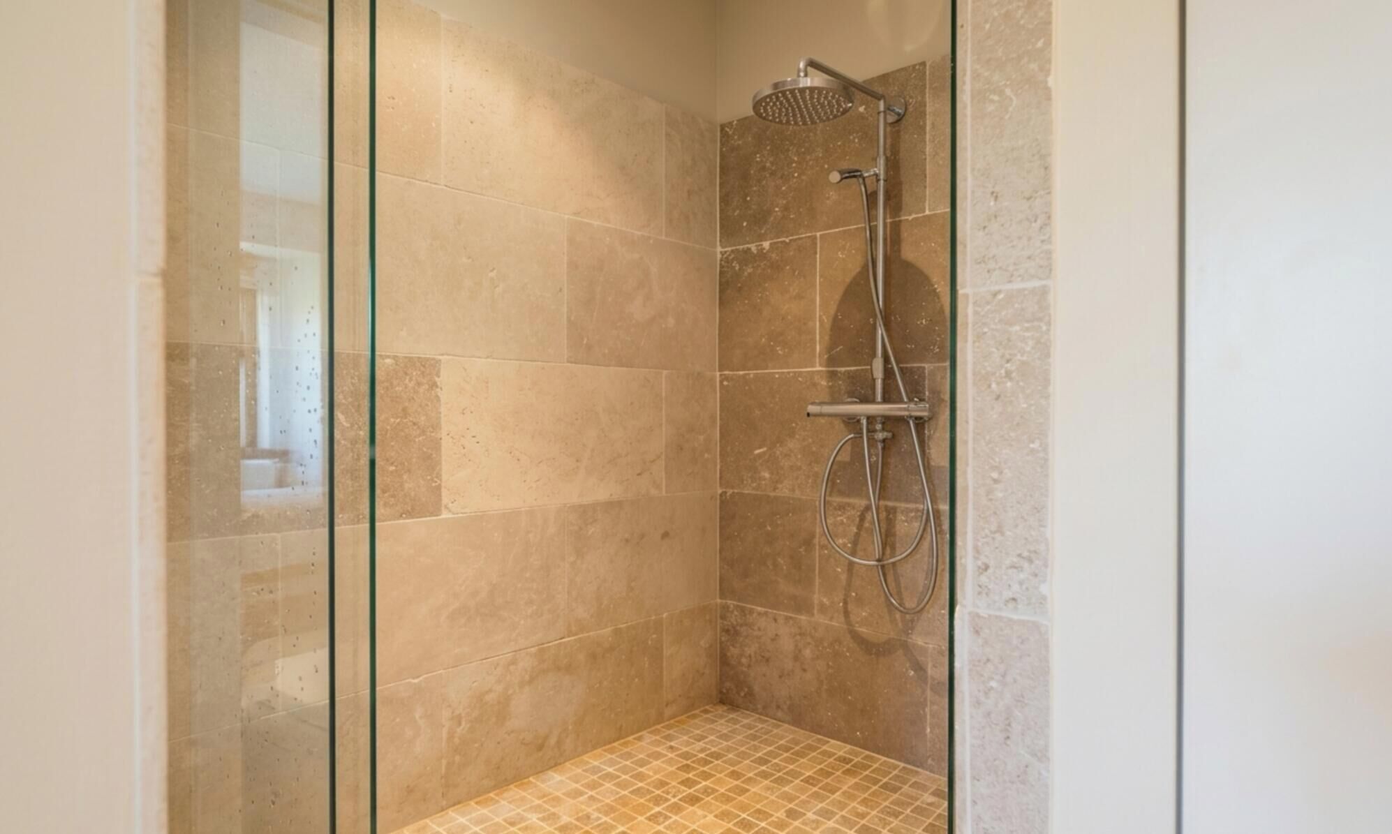 Een douchecabine met een glazen wand, een handdouche en een muurbadkraan.