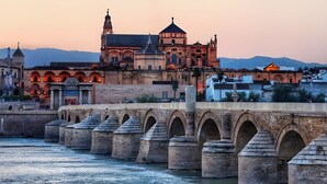 A stone bridge with arches over a river, leading to a historic building with multiple domes and a clock tower.