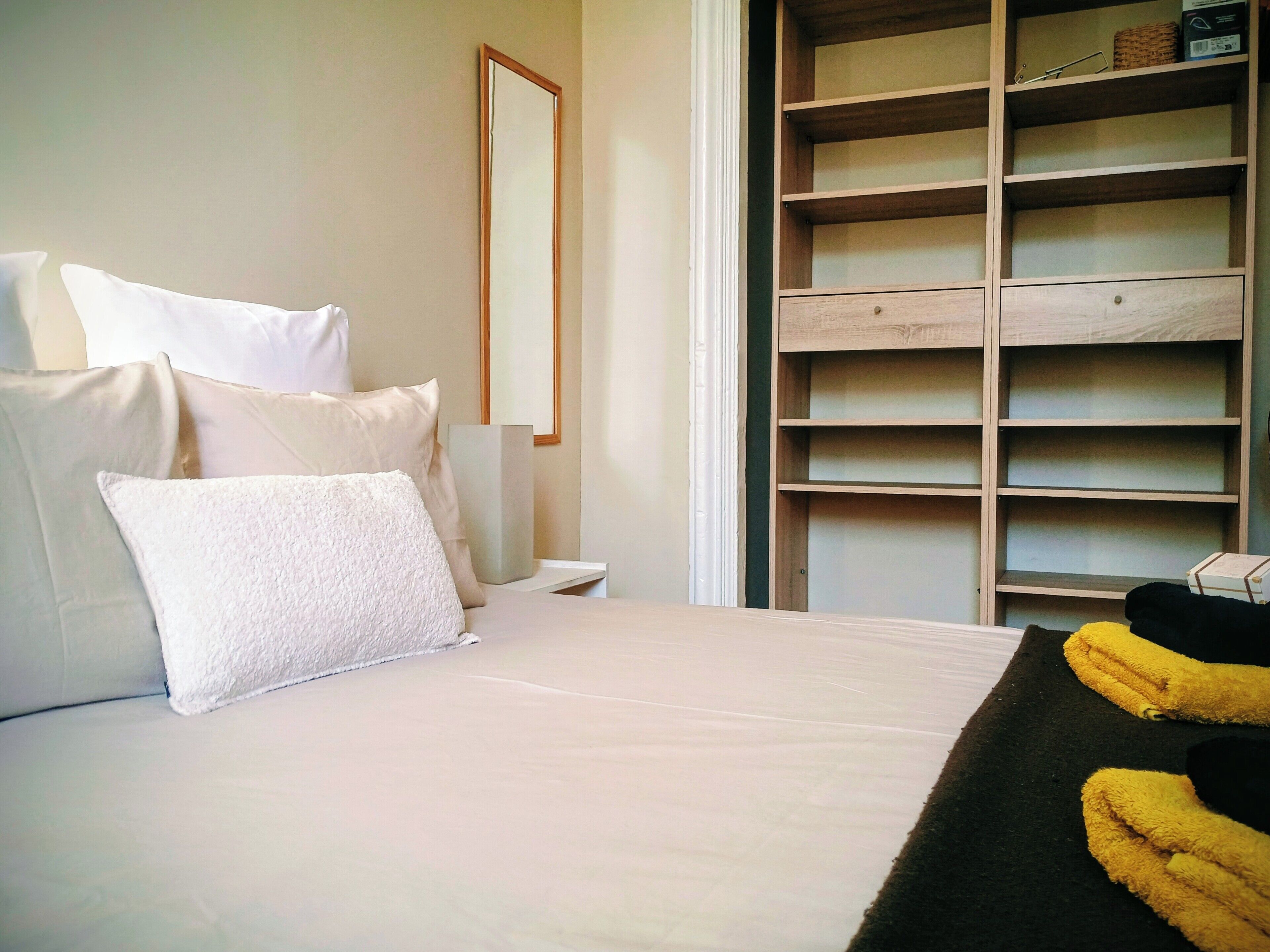 A bedroom with a bed, pillows, a bedside table, a mirror, and a wooden wardrobe with shelves.