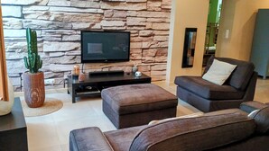 A living room with a stone wall, a flat-screen TV, a cactus, and a sofa. - L'Isle Contemporaine - Spacious 74 M2 Clim Parking Downtown 2 bedrooms (L'Isle-sur-la-Sorgue)