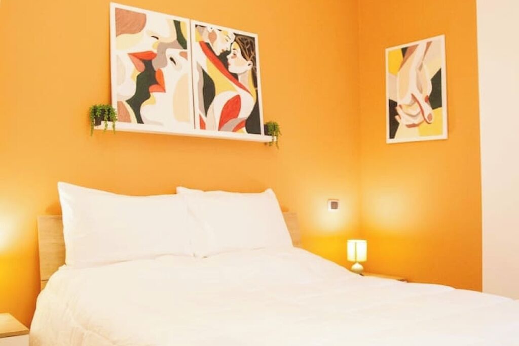 Une chambre d’hôtel avec un grand lit, une table de chevet, une lampe fixée au mur et deux tableaux abstraits.
