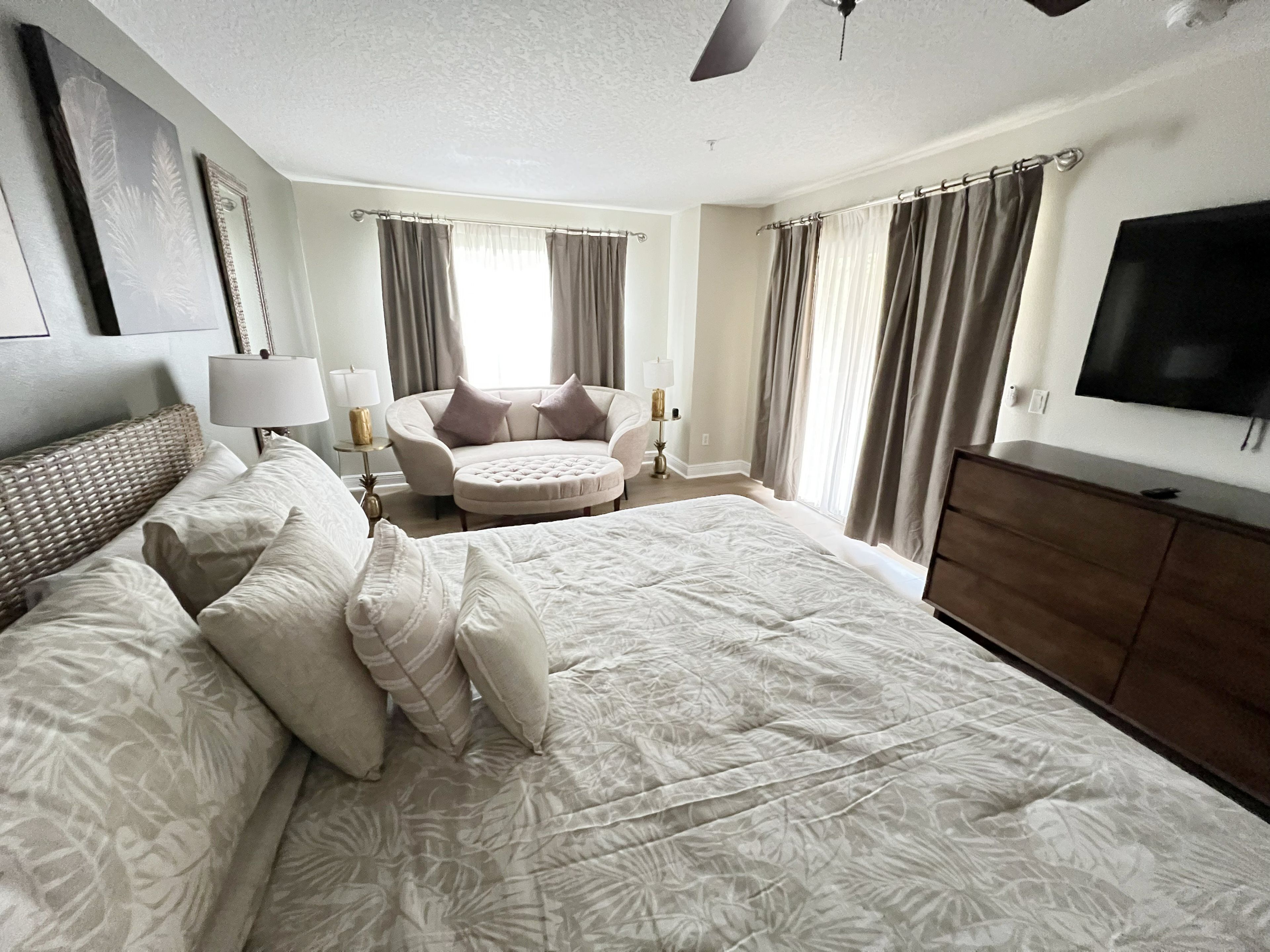 A spacious hotel room with a large bed, a round seating area, a TV, and a ceiling fan.