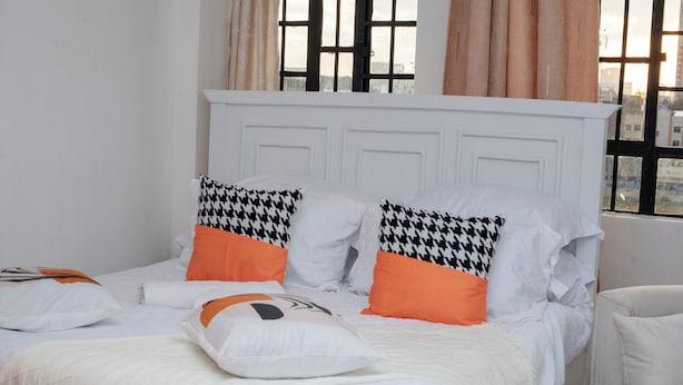A neatly made bed with white bedding and orange and black patterned pillows, a white throw blanket, and a white chair beside the bed. - Couple Friendly Studio Apartment In Pangani (Nairobi)