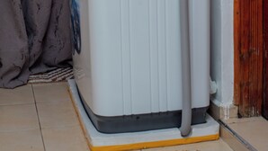 A white portable heater with a hose, placed on a yellow platform in a room with a tiled floor and a wooden door. - Couple Friendly Studio Apartment In Pangani (Nairobi)