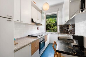 Une cuisine moderne avec des meubles blancs, un four encastré, une cuisinière à gaz, une machine à café et une fenêtre donnant sur un paysage verdoyant.