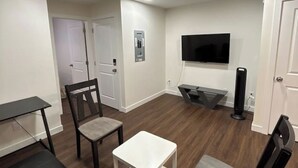 Una habitación con un televisor de pantalla plana montado en la pared, un pequeño escritorio blanco, una silla de madera oscura y un saco de boxeo negro.